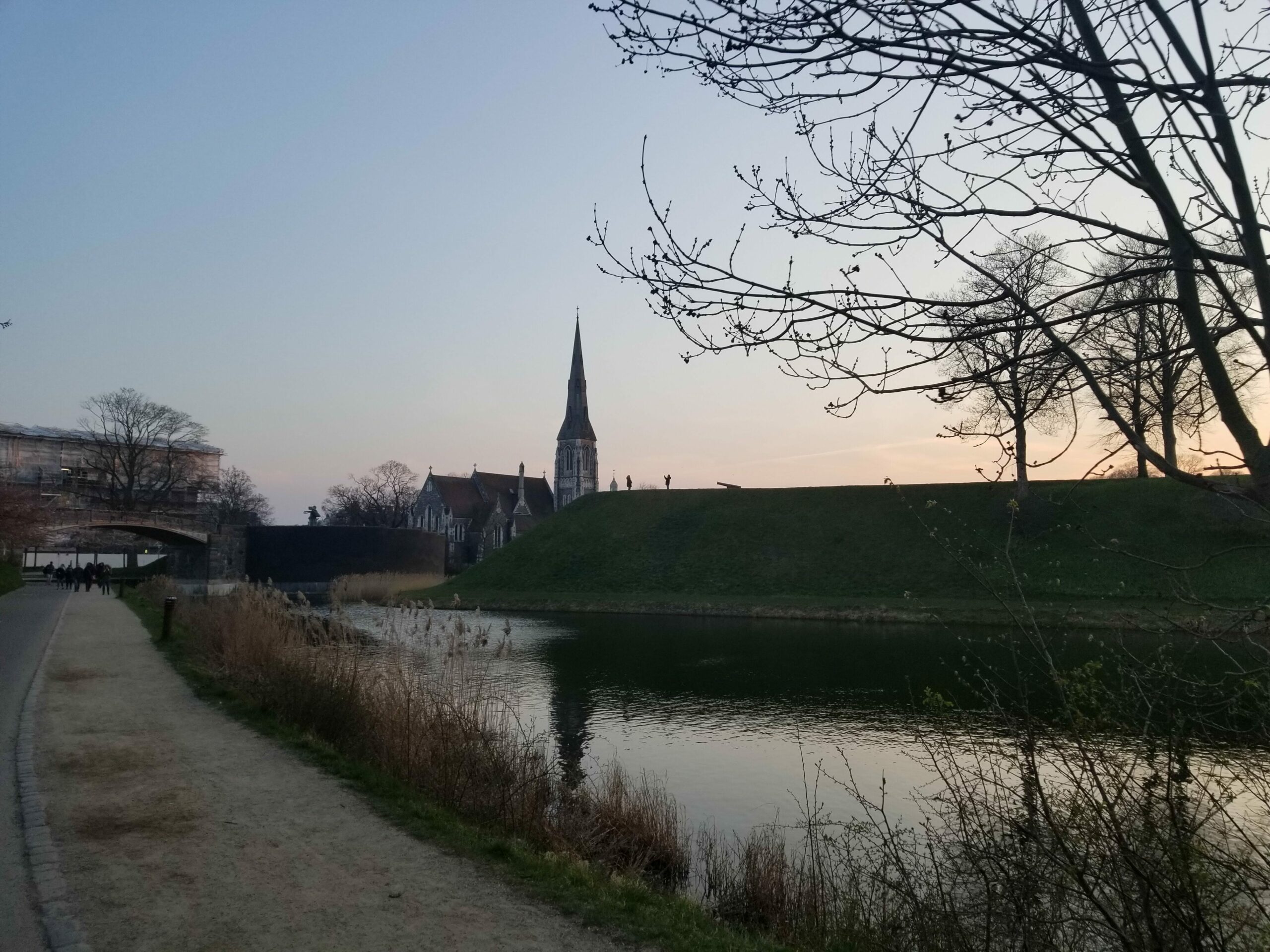 A spire rises over a sunset-lit moat and bike path.