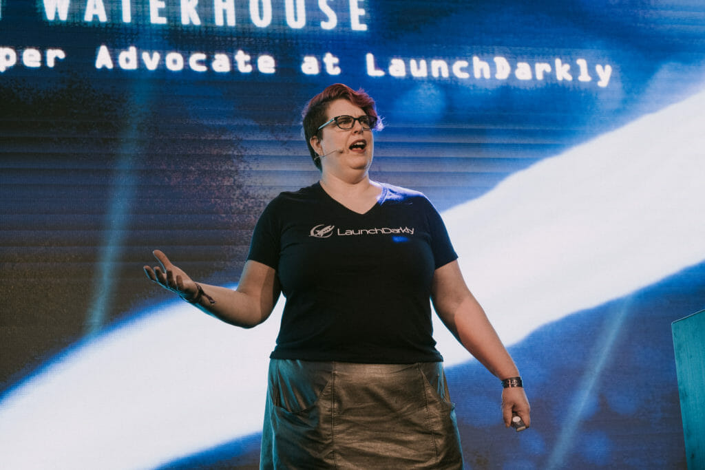 A white woman in mid-speech. She is wearing a headset mic, a company t-shirt, and a silver skirt.