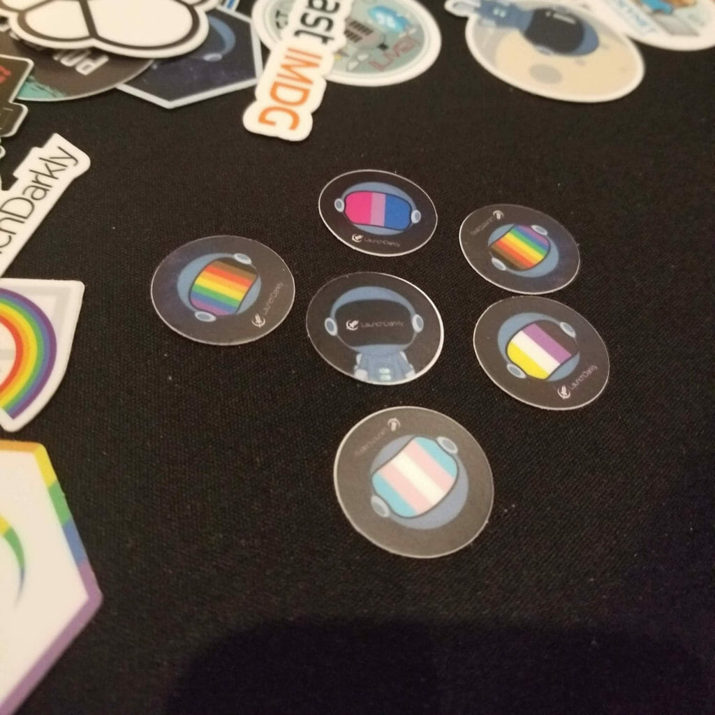A set of six 1-inch round stickers with a space helmet that has flags for trans, bi, and non-binary pride.