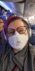 A pink-haired white woman on a plane. She is wearing an N95 mask and over-the-ear headphones. Her hair is braided across her hairline, and her glasses are burgundy.