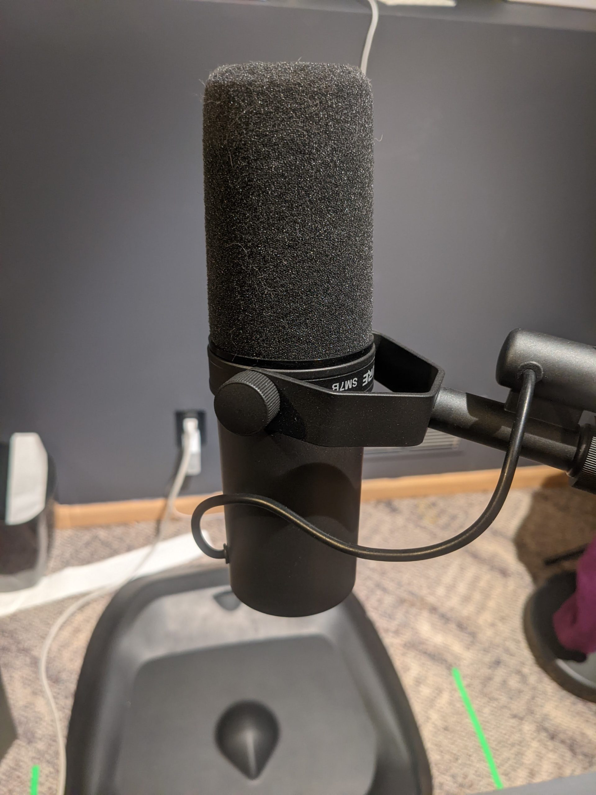 A Shure SM7-B microphone on a stand. The background is a standing mat and a wall painted grey