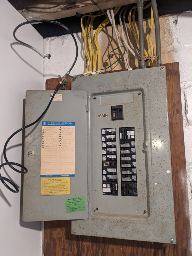 An electrical panel, which is a grey metal box with a putative description of the circuit destinations on the inside of a door standing open, and 20 circuit breakers on the right side of the box. A bunch of various colored wires run up from the box. The box is a little bit rust-speckled
