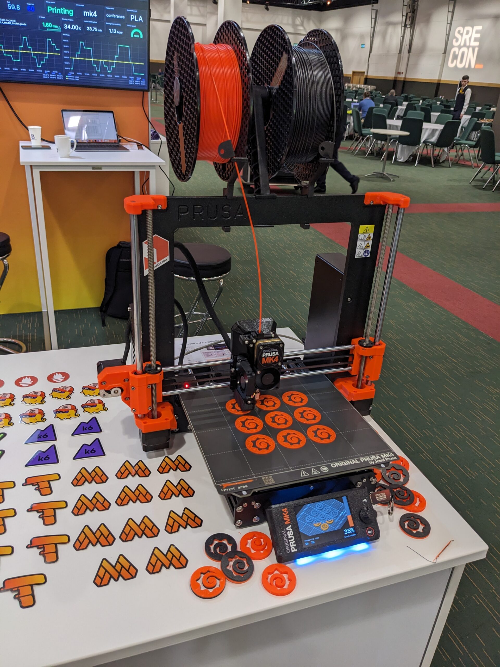 A 3D printer at a trade show booth. It is printing orange spirals that are the Prometheus logo
