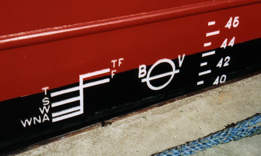 The side of a ship. The top part is painted red, and the bottom is black. Painted on it is a white diagram with marking starting at TF at the top, then F, T, S, W, and WNA. There is also a circle with a horizontal line through it slightly below the point where the paint changes.