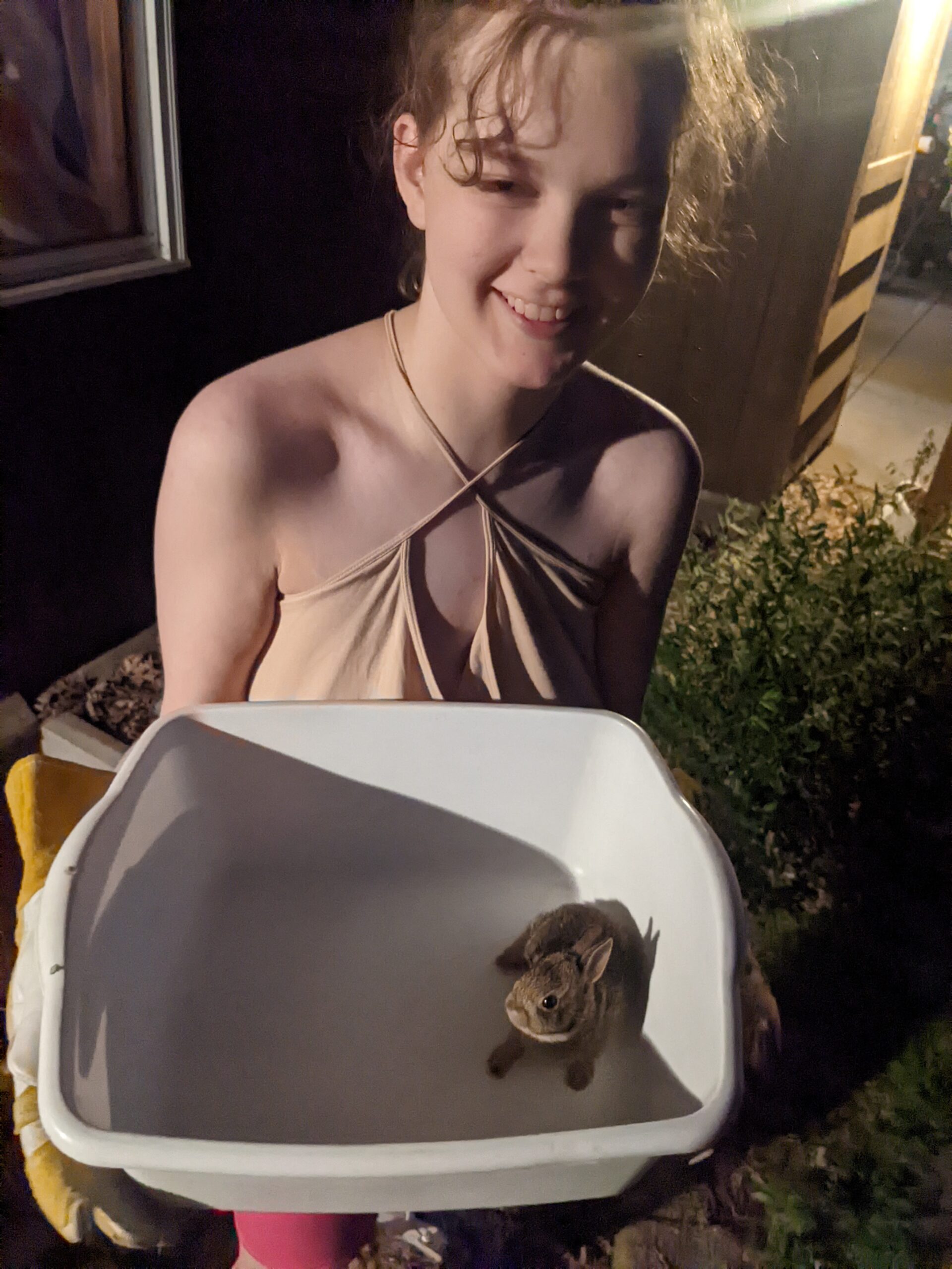 A young person with curly hair is holding a dish pan with a tiny bunny in it. The picture is taken outside at night.