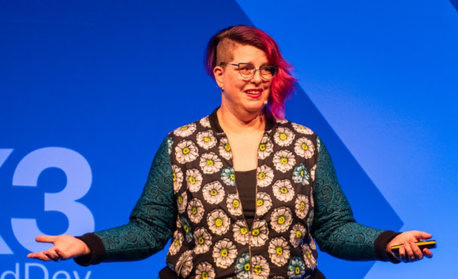 A white woman with pink hair gestures widely as she stands on stage.